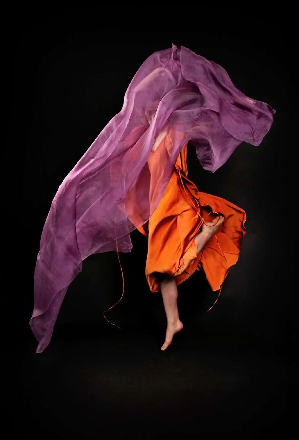 Modelo girando con un vestido naranja y una tela violeta suspendida, sobre fondo negro