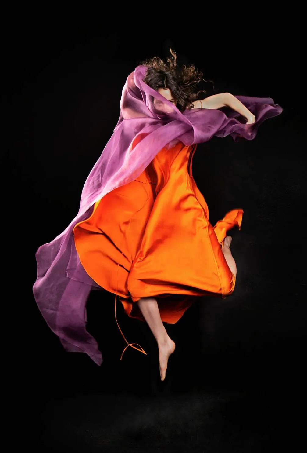 Modelo saltando con un vestido naranja y una tela violeta al viento, sobre fondo negro