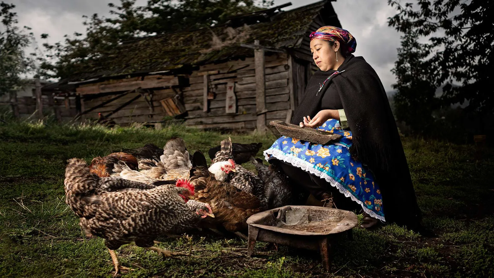 Mujer mapuche sentada al aire libre junto a una casa de madera