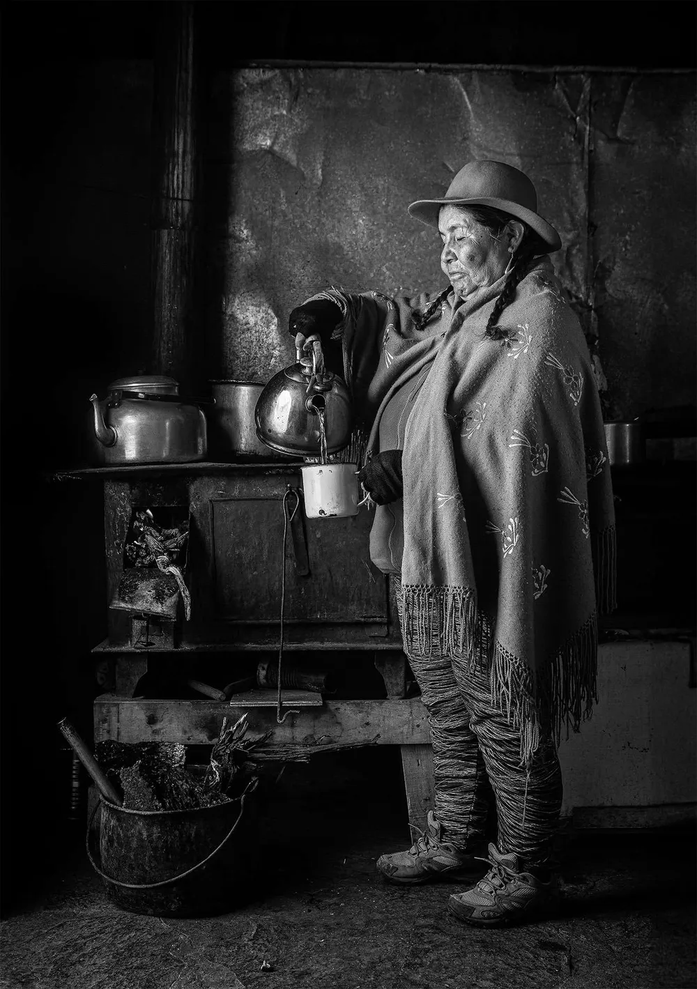Mujer aymara de pie junto a una cocina a leña, en blanco y negro